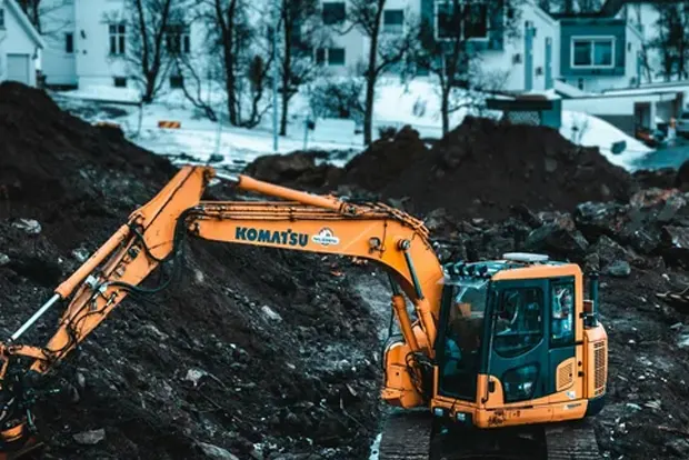 Yellow excavator digging in a snowy construction site with trees and houses.